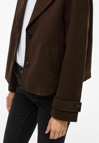 Chaqueta marrón con botones y cintas en los puños, llevada sobre una camisa blanca y pantalones negros, recortada a la altura de la cintura mostrando el brazo izquierdo y la mano relajada.