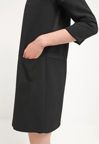 Robe noire avec des manches trois-quarts, une coupe ample et une poche à l'avant. Texture lisse avec un design minimaliste et une longueur au genou.