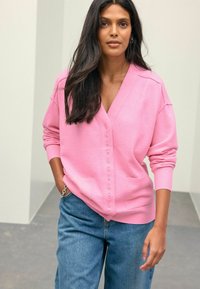 Rosa Strickjacke mit V-Ausschnitt, Knopfleiste und Seitentaschen, aus weichem Material. Über blauer Denim getragen, mit gerippten Bündchen und Saum.