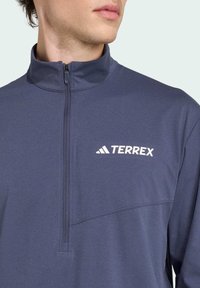 Námornícky modrý pulóver s dlhým rukávom a polovičným zipsom, s vysokým golierom a bielym logom Terrex na hrudi. Hladká, ľahká tkanina.