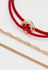 Collier en corde tressée rouge avec un détail en boucle dorée, accompagné de deux chaînes dorées : une chaîne à maillons plats et une chaîne serpent.