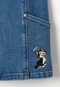 Denimstoff in Hellblau mit einer Tasche. Die Vorderseite zeigt eine gestickte schwarz-weiße Cartoon-Katze mit dem Text "HEY!" in einer verspielten Schriftart.