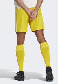 Geel atletiekshorts met witte zijkanten, gecombineerd met bijpassende gele kniekousen en zwarte voetbalschoenen.
