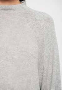 Harmaa neulepaita korkealla kauluksella, jossa on raglan-hihat ja pehmeä tekstuuri. Saumayksityiskohdat tuovat hienovaraista visuaalista mielenkiintoa.