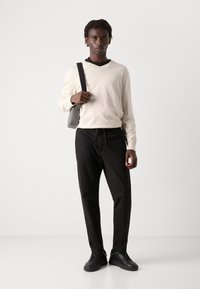Crème trui met lange mouwen en een v-hals, zwarte drawstring broek, zwarte sneakers en een grijze schoudertas tegen een effen achtergrond.