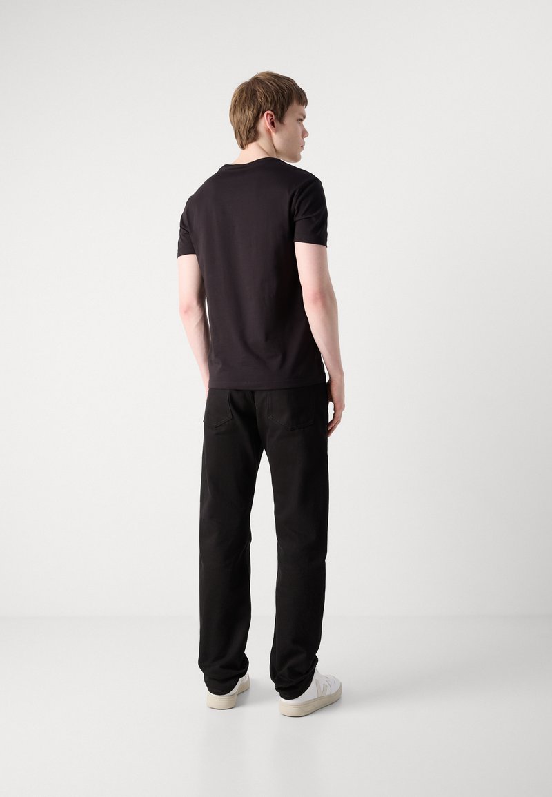 Joven de pie, de espaldas, vestido con una camiseta negra, pantalones negros y zapatillas blancas sobre un fondo blanco liso.