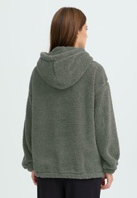 Grüne, flauschige Kapuzenjacke aus plüschigem, strukturiertem Material. Verfügt über eine geräumige Kapuze und einen lockeren Schnitt mit gerippten Bündchen, die bis zur Mitte des Oberschenkels reichen.