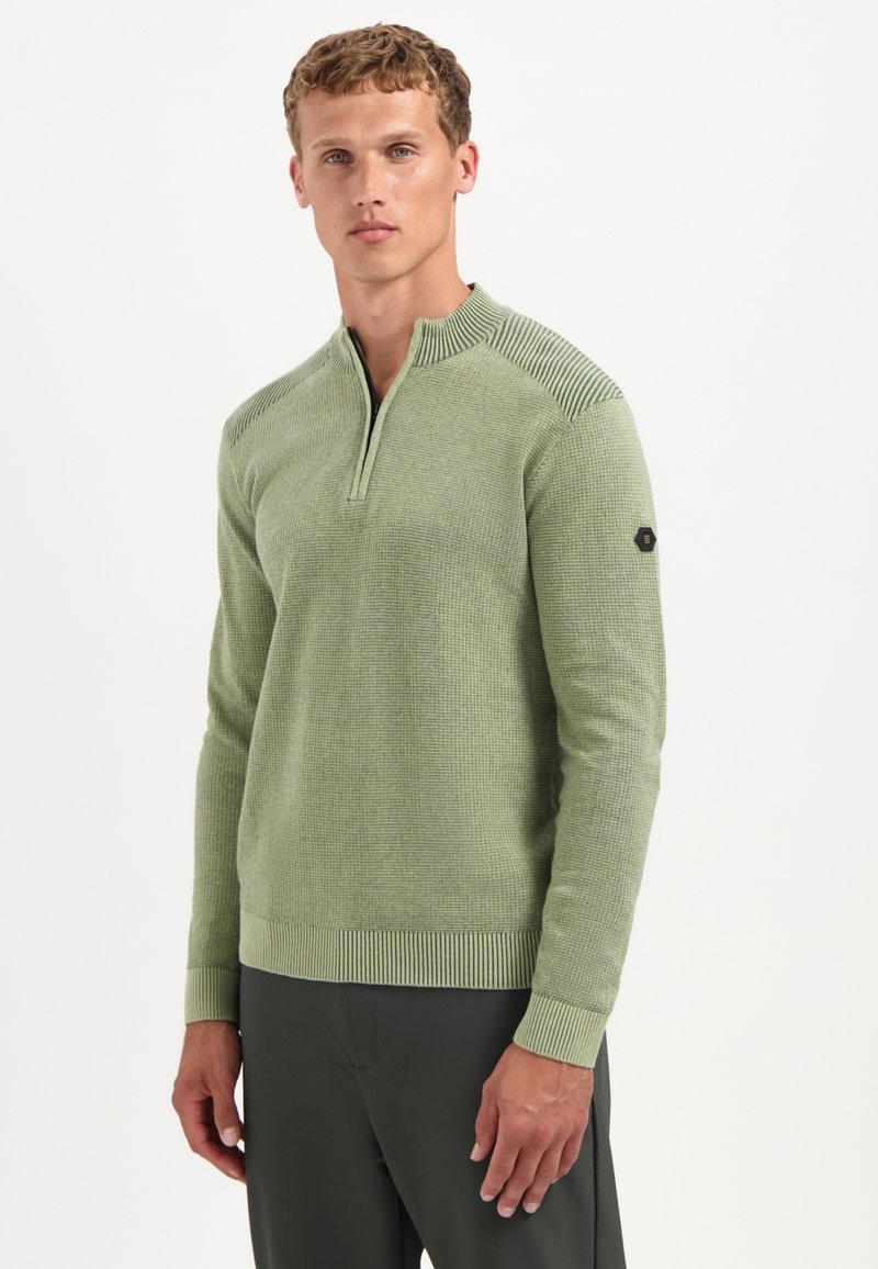 Man staat met het gezicht naar voren en draagt een lichtgroene textuur half-zip trui en donkere broek tegen een witte achtergrond.