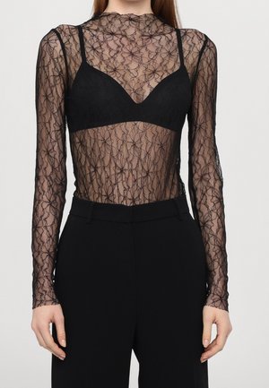 Woman wearing long-sleeve sheer black floral lace top over black bralette and black high-waisted trousers, hands relaxed at sides.