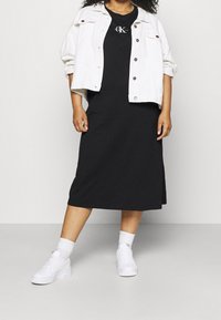 Osoba z kręconymi włosami nosząca biały krótki żakiet na czarnej sukience midi od Calvina Kleina, w białych skarpetkach i białych sneakersach.