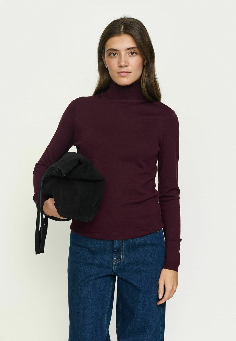 Jeune femme aux longs cheveux bruns porte un col roulé violet foncé et un jean bleu, tenant un sac noir plié sous le bras.