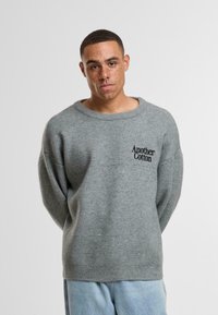 Maglione grigio lavorato a maglia con vestibilità ampia, scollatura larga e logo testuale nero "Another Cotton." Indossato con pantaloni in denim azzurro chiaro.