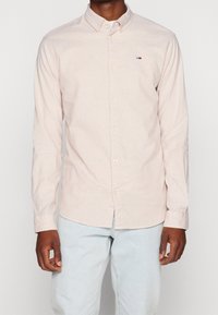 Man in een lichtroze, knooploze lange mouwen overhemd met een klein vlag-logo op de borst en lichtblauwe spijkerbroek.