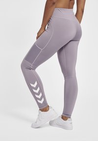 Lilla højtaljede leggings med en glat tekstur, sidelommer og et hvidt chevronmønster på det ydre ben. Båret med hvide sneakers.