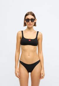 Črni rebrasti bikini komplet s kvadratnim izrezom in širokimi trakovi, združen z ujemajočimi se nizkimi hlačkami. Okrasen z majhnim rdečim logotipom.