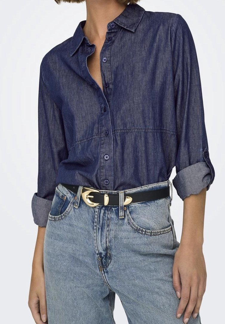 Woman wearing dark blue button-up denim shirt with rolled sleeves, light blue jeans, and black belt with gold buckle.