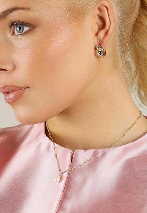 Guldringar med klara stenar; smal silverkedja med en ensam pärlhängsmycke. Slätt, ljusrosa tygskjorta.