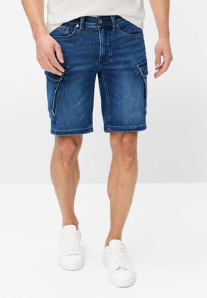 Man draagt donkerblauwe denim cargoshorts van middellange lengte met zijflapkanten en witte sneakers, staand tegen een effen achtergrond.