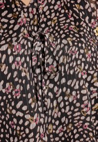 Schwarze Bluse aus Stoff mit einem Bindeband am Ausschnitt, mit einem Muster aus pinken, weißen und goldenen Punkten, strukturierten Oberfläche.