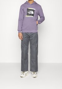 Sudadera morada con un logo frontal, pantalones cargo grises con costuras reforzadas y zapatillas blancas con detalles verdes. Conjunto casual en general.