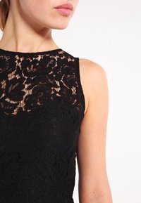 Gros plan sur une femme portant une robe en dentelle noire sans manches avec des motifs floraux et un tissu transparent sur le haut de la poitrine.