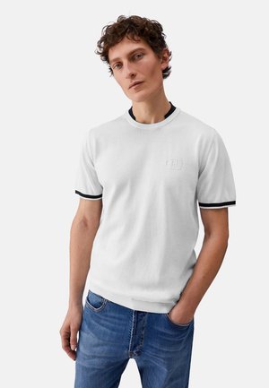 Giovane uomo con capelli ricci che indossa una camicia bianca a maniche corte con bordo nero e jeans blu, in piedi con una mano in tasca su uno sfondo semplice.