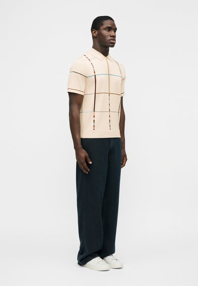 Modèle masculin portant un polo beige à manches courtes avec des rayures verticales multicolores, un pantalon large foncé et des baskets blanches, debout de profil.
