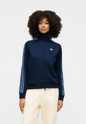 Vrouw met krullend haar, gekleed in een marineblauwe jas met rits en lichtblauwe strepen op de mouwen en beige broek, staand tegen een effen achtergrond.