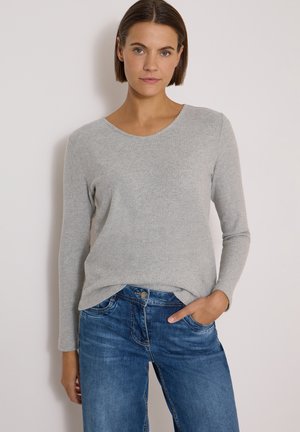 Jeune femme aux cheveux courts et bruns portant un pull gris clair à manches longues et un jean bleu, une main dans la poche, sur un fond uni.