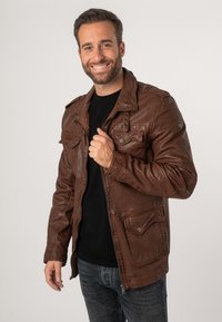 Braune Lederjacke mit einer robusten Textur, Reißverschluss vorne und mehreren Fronttaschen. Mit Schulterklappen und Metall Druckknöpfen.