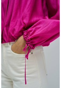 Mano in tasca, indossa pantaloni bianchi e una blusa magenta a maniche lunghe con polsini arricciati e dettaglio annodato al polso.