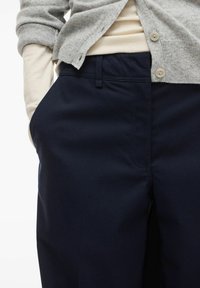 Pantalon bleu marine sur mesure avec une texture lisse. Dispose d'une poche avant et de passants pour ceinture, accompagné d'un cardigan gris clair porté par-dessus.