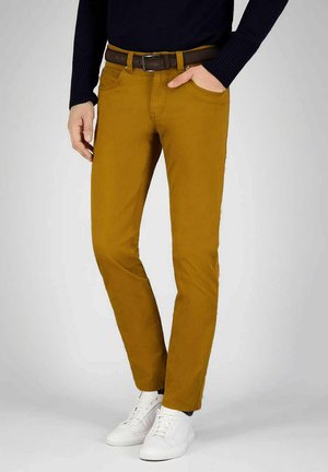 Man die mosterdgele slim-fit broek draagt, met een bruine riem, zwarte trui, witte sneakers, met één hand in zijn zak tegen een effen achtergrond.