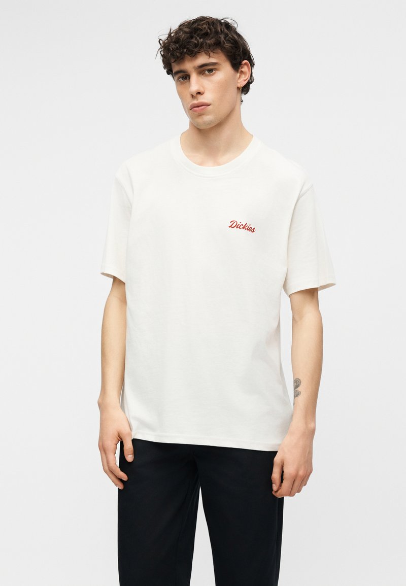 Jeune homme aux cheveux bouclés portant un t-shirt blanc à manches courtes avec le logo rouge "Dickies" et un pantalon noir, debout devant un fond uni.