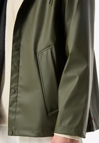 Primer plano de una persona con chaqueta impermeable verde oliva con botones a presión y bolsillo lateral inclinado, sobre un suéter de punto blanco.