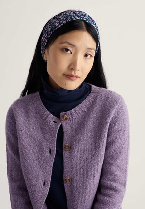 Alyvinės spalvos megztinis su apvalia apykakle, mediniais mygtukais ir tekstūruota medžiaga, dėvimas ant tamsios rolkės. Gėlėtu raštu išmarginta galvos juosta.