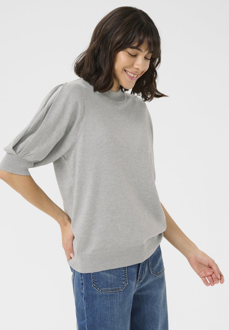 Pull en tricot gris clair avec un col haut, des manches courtes bouffantes et un ourlet côtelé. Associé à un jean droit bleu foncé.