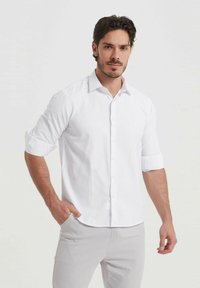 Chemise blanche à col déployé, avec des manches retroussées et un tissu texturé. Portée avec un pantalon gris clair, mettant en valeur une coupe ajustée.