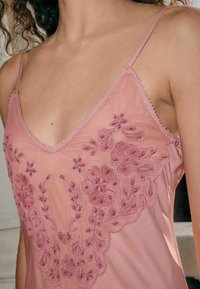 Camisola in seta rosa con ricami floreali intricati e perline; spalline regolabili e scollatura a V profonda. Tessuto liscio, design elegante.