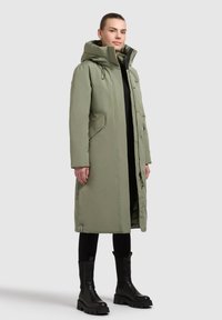 Geacă lungă de tip puffer în verde măsliniu, cu glugă, având două buzunare laterale și fermoar în față. Purtați-o peste pantaloni negri și cizme cu talpă groasă negre.
