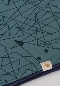 Textile mat with a teal background, featuring a geometric pattern of dark lines and triangles. Includes a small tag with a light design.