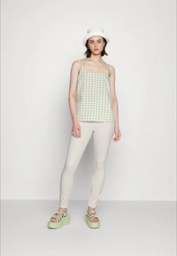 Jovem mulher a usar um chapéu de balde branco, uma blusa sem mangas em xadrez verde, calças bege e sandálias de plataforma verde claro, de pé contra um fundo simples.