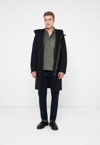 Zwarte lange jas met capuchon, knopen en ritsen over een groene knoopshirt, marineblauwe broek en zwarte schoenen. Eenvoudig, modern ontwerp.