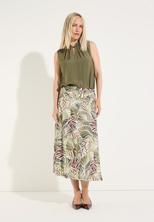 Femme portant un haut sans manches couleur olive et une longue jupe à motifs avec des feuilles vertes, blanches et rouges, associée à des chaussures pointues foncées.