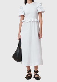 Vestido maxi blanco con bodice fruncido, cintura con volantes y mangas cortas abullonadas. Bolso negro con acabado texturizado y acentos dorados; sandalias planas con hebillas.