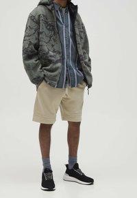 Kurtka polarowa z kapturem w kolorze oliwkowym z abstrakcyjnym wzorem, koszula z paskami na guziki, beżowe szorty, czarne trampki z białymi podeszwami, szare skarpetki.