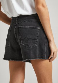Pantalones cortos de mezclilla negra con dobladillo deshilachado, dos bolsillos traseros con costuras sutiles y un acabado desgastado en la textura de la tela.