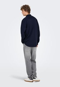 Marineblå langermet skjorte med krage, løs passform, kombinert med lysegrå jeans og hvite joggesko med mørke detaljer; innendørs setting.