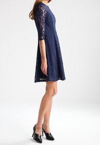 Marineblauwe kanten jurk met een aansluitende bovenkant en een uitlopende rok. Heeft driekwart mouwen en een gegolfde zoom.