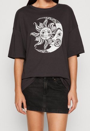 Person trägt ein schwarzes oversized T-Shirt mit einer weißen Sonne- und Mond-Gesichts-Grafik, kombiniert mit einem schwarzen Denim-Minirock.
